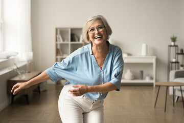 Joyful happy retired elderly woman in glasses having fun at home, dancing to music in modern...