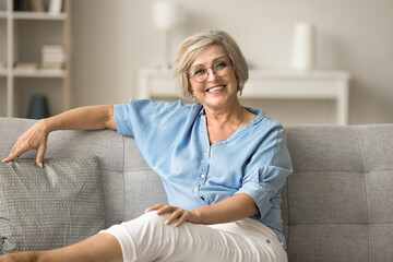 Happy pretty mature old woman in glasses resting on soft couch, looking at camera with toothy...