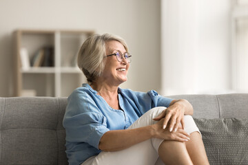Positive blonde older mature woman in stylish glasses sitting on home couch, looking away, smiling,...