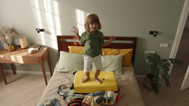 Active child boy jumping on suitcase. Bag luggage with poorly packed clothes sticking out, little boy tourist traveler jumping on baggage suitcase raising hands high up. Happy child jumps with delight