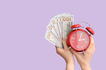 Female hands holding alarm clock and money on lilac background