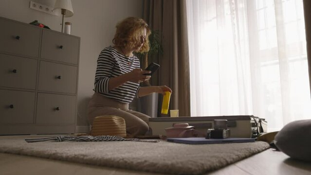 Woman packs suitcase with phone in hand. Sitting on floor female tourist traveler packs luggage puts clothes in suitcase in organized manner checking list in smartphone. Calm leisurely luggage packing