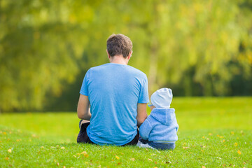 Young adult father and baby boy sitting together on green grass at park. Spending time together in...