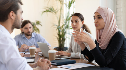 Multicultural businesspeople negotiating gathered together at office group meeting led by businesswoman middle eastern ethnicity leader in hijab, talking expressing opinion speak with european partner
