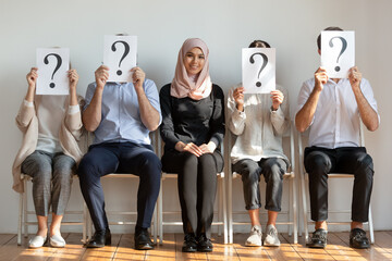 Fototapeta na wymiar Diverse people sit on chairs hiding faces behind questions marks waiting job interview. Successful applicant middle eastern ethnicity arabian female got position in international company, hr concept