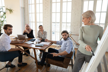 Arabian, mixed-race and european diverse company staff members listen mature female 60s business coach explain data on flip chart share knowledge helpful info during meeting or workshop at board room