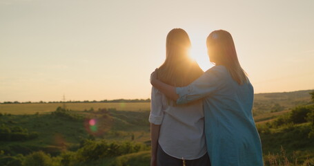 Mom hugs her teenage daughter, watching the sunset over a picturesque valley. Back view