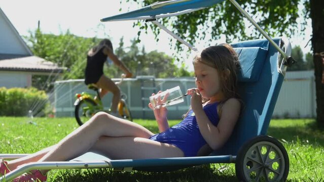 Little girl in blue swimsuit takes a glass with a drink sitting in a deck chair