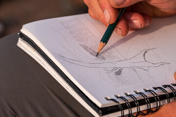 making art outdoors: man's hands  drawing on a sketch book with a pencil close up	