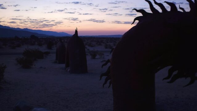 Aerial: Drone Panning Shot Of Metallic Sand Serpent Sculpture Amidst Plants Near Mountains Against Cloudy Sky At Dawn - Borrego Springs, California