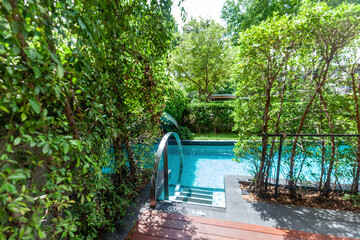 Swimming pool surrounded by trees and bushes. Outdoor oasis, relaxation, summer, vacation. Close-up of blue swimming pool with ladder. Luxury swimming pool with trees and sundeck