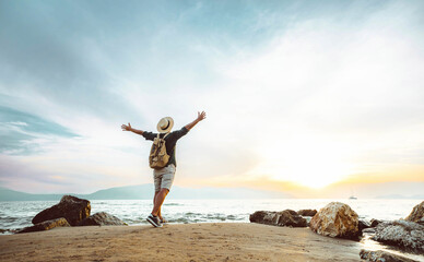 Happy man with backpack standing with arms up at the beach - Delightful tourist enjoying summer vacation by the seaside - Traveling life style and well being concept - 651643437