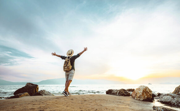 Happy man with backpack standing with arms up at the beach - Delightful tourist enjoying summer vacation by the seaside - Traveling life style and well being concept