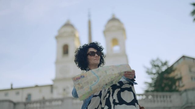 Attractive curly hair latin businesswoman sits on a staircase of the Spanish steps in Rome, italy holding city map and hand pointing at landmarks Background view of church Trinit&agrave; dei Monti