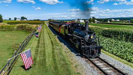 An Aerial View of a Steam Passenger Train Approaching, on a single Track With a Fence and America Flags Blowing in the Wind, on a Summer Day - obrazy, fototapety, plakaty