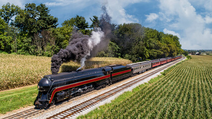 An Aerial Front View of a Restored Antique Steam Passenger Train, Traveling Thru the Countryside, Passing Corn Fields and Blowing Smoke on a Sunny Summer Day - obrazy, fototapety, plakaty