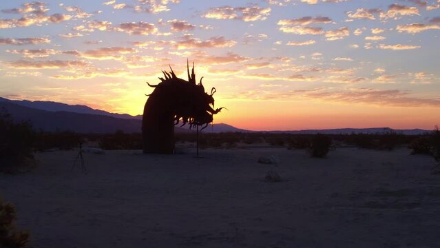 Aerial: Drone Panning Shot Of Metallic Sculpture Amidst Plants By Road In Desert Against Cloudy Sky During Sunrise - Borrego Springs, California