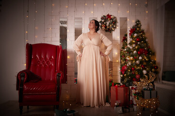 Owerweight elegant Woman at Christmas room. Fat plumb pretty girl in a beautiful dress for a holiday. Popular buxom female model posing alone in New year sudio