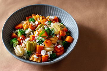 Autumn pasta salad with roasted pumpkin, broccoli, feta cheese on dark wooden background, vivid colors fall season salad, autumn food concept, top view
