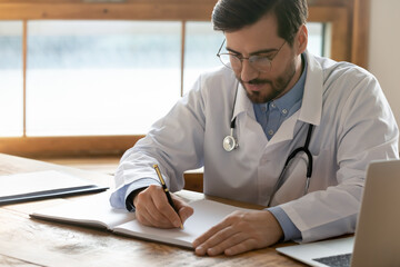 Focused doctor wearing glasses writing illness history, sitting at workplace in hospital, serious man gp therapist wearing white coat uniform with stethoscope filling patient card, paperwork - obrazy, fototapety, plakaty