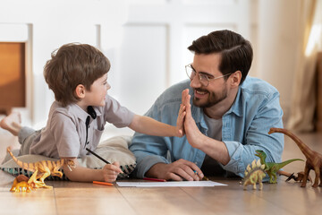 Happy father wearing glasses and little son giving high five, playing with toys, drawing colorful pencils, lying on warm wooden floor with underfloor heating, family having fun at home