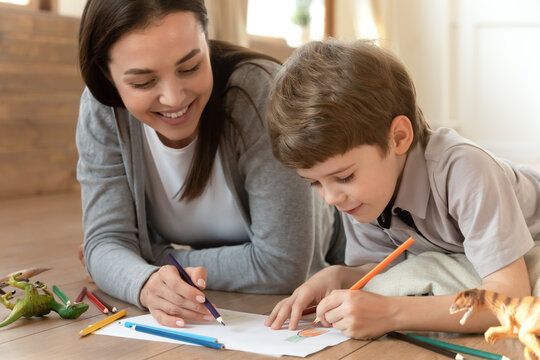 Close up happy mother and little son drawing colorful pencils on paper sheet together, having fun, lying on warm wooden floor with underfloor heating, family enjoying creative activity at home