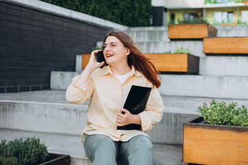 Elegant young 30s woman formally dressed, sitting on the bench and holding a folder with documents. Businesswoman talking on cell phone, making call and standing on modern city street.