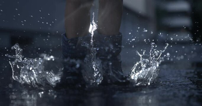 Nostalgic scene of child wearing rain boots jumping in the air in water puddle having fun in ultra slow-motion captured in high-speed camera