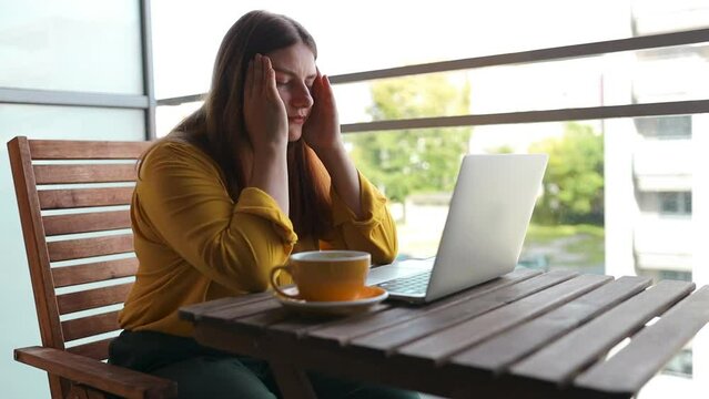 Young Caucasian businesswoman with headache while working with laptop in modern cafe. Stressed and depressed girl, touching her head, feeling pain, sitting at a wooden table in a cafe.High quality