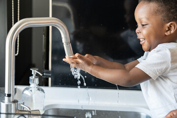 Cute little smiling African-American boy washing hands in a kitchen sink at home