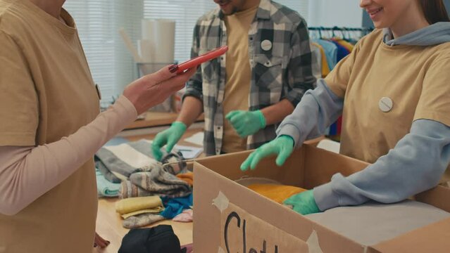 Medium Shot Of Small Group Of Volunteers Collecting Clothes On Box For Those In Need While Woman Recording Video On Mobile Phone For Donation Centre Blog