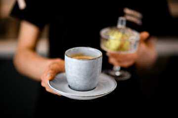 Barista holds cup of gentle cappuccino with thick foam and crystal vase with tasty sweet macaroon