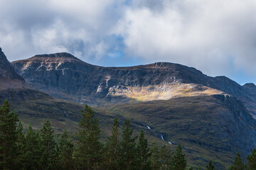 Steep mountain view. Sun lighting part of the mountain. Evergreen forest on foreground. Northern Norway.