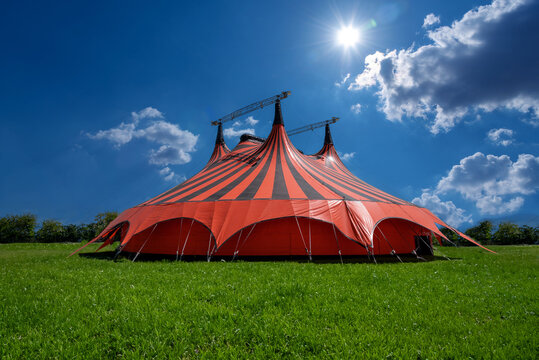 Red and black circus tent in green meadow with blue sky with clouds and sunshine