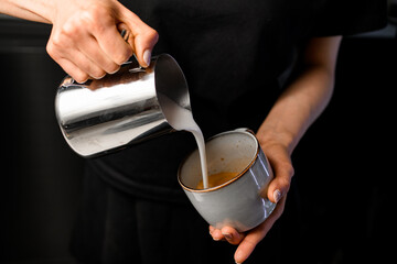 Cropped shot of female barista pouring milk from metalllc pitcher into cup with coffee