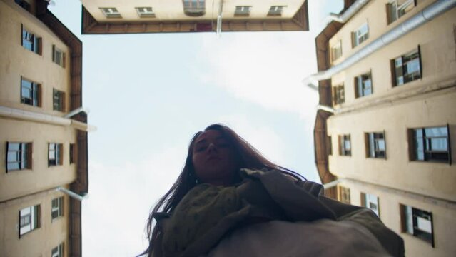 Low-angle of a young caucasian girl with long hair looking down at the camera. Brown residental houses with old windows and drainage pipes in a yard-well and blue cloudy skies in St. Petersburg around
