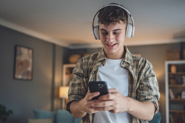 Portrait of teenage boy stand at home use headphones ans smartphone to play music or watch video...