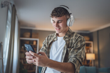 Portrait of teenage boy stand at home use headphones ans smartphone to play music or watch video...