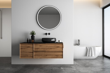 Bright bathroom interior with white tones wall, concrete floor, wooden vanity with black sink and oval mirror, white bathtub, panoramic windows. - obrazy, fototapety, plakaty