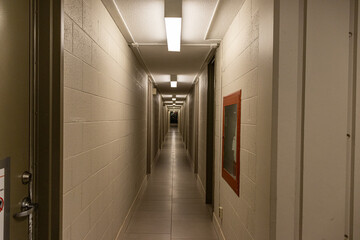 A long, narrow hallway with closed gray doors and a red fire extinguisher - white walls and tiled floor - fluorescent lights overhead. Taken in Toronto, Canada.