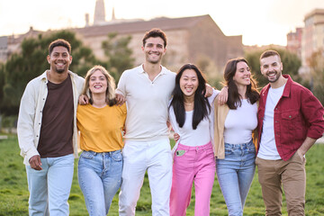 Happy young beautiful people enjoying summer sunny day spending time outdoors and having fun together. Joyful and smiling multiracial friends hugging standing in city park. Diverse group portrait.