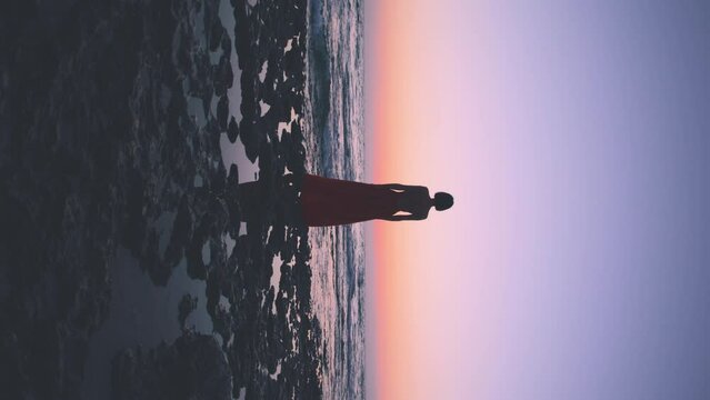 vertical shot. A young woman in a summer dress stands on a rock during sunset and looks at the sea. freedom and beauty.
