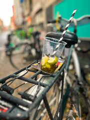 Thirsty Bicycle. Empty plastic drink cup with striped drinking straw rests on the back of a parked bicycle. There are limes at the bottom of the cup. The bicycle is parked in a city alley in Cologne.