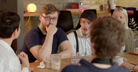 Group discussion at the table. Woman speaker speaks information. A man touches his glasses, an obsessive movement. A bearded man listens with his mouth open.