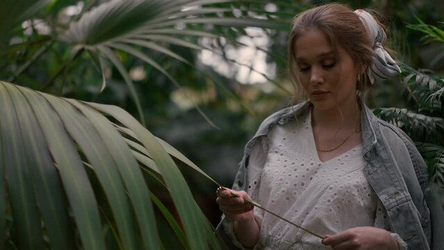Romantic blond woman touching palm leaf in botanical garden portrait