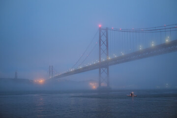 Fototapeta premium View of 25 de Abril Bridge famous tourist landmark of Lisbon connecting Lisboa and Almada in heavy fog mist wtih yacht boats passing under. Lisbon, Portugal