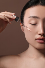 Fototapeta premium Beautiful young woman applying cosmetic serum onto her face on brown background, closeup