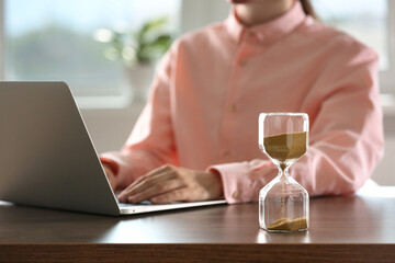 Hourglass with flowing sand on wooden table, selective focus. Man using laptop indoors
