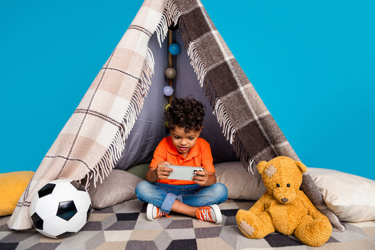 Photo of cute bpy little gamer using smart phone playing in diy tent isolated blue color background