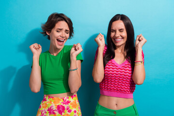 Photo of excited lucky ladies wear colorful clothes rising fists screaming yeah isolated blue color...